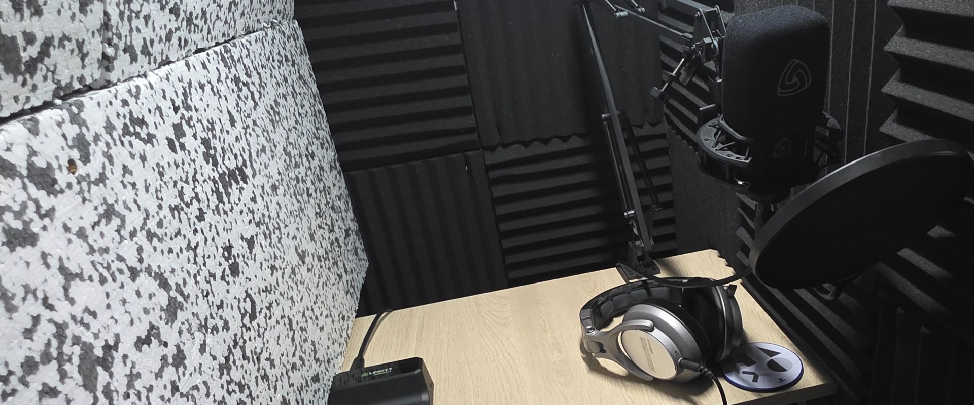 A small recording booth with a Lewitt microphone with a pop filter in front of it, attached to an arm on the right. There is a pair of Shure headphones and a PlayStation coaster underneath the mic, and part of a Lewitt audio interface is on the left.
